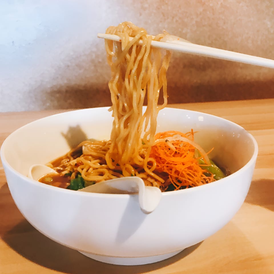 Chopsticks lifting ramen noodles from a white bowl with shredded carrots and green vegetables.